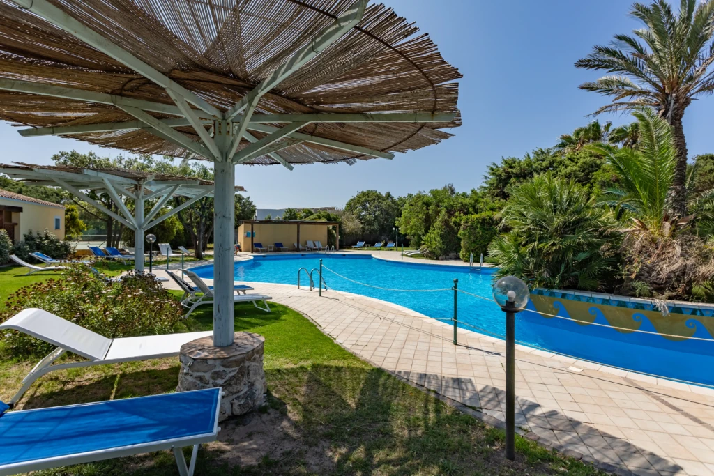 Piscina dell'Hotel Luci del Faro con lettini blu, ombrelloni in paglia e giardino mediterraneo a Calasetta
