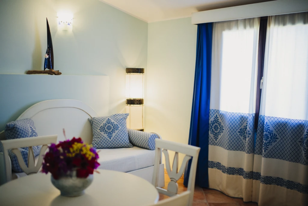 Camera matrimoniale con balcone vista mare, letti bianchi, tende blu e tavolo con fiori colorati all'Hotel Luci del Faro