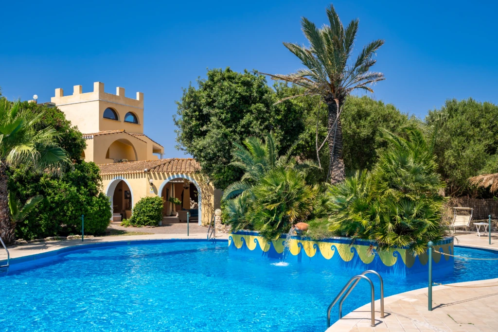 Piscina esterna dell'Hotel Luci del Faro circondata da palme e giardino con edificio in stile mediterraneo