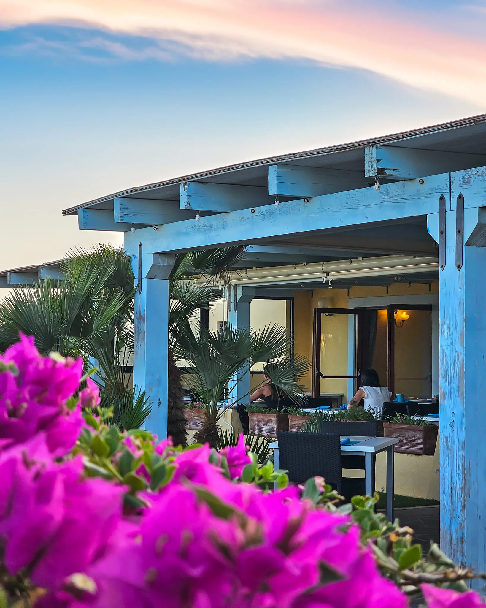 Pergola blu con area pranzo esterna al tramonto, fiori fucsia in primo piano all'Hotel Luci del Faro