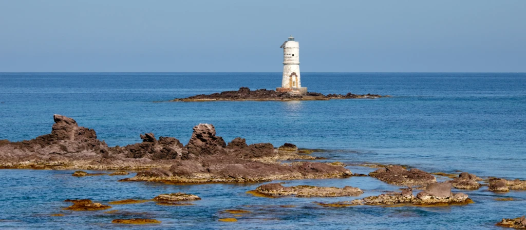 Faro bianco su isolotto roccioso con scogli in primo piano e mare azzurro calmo
