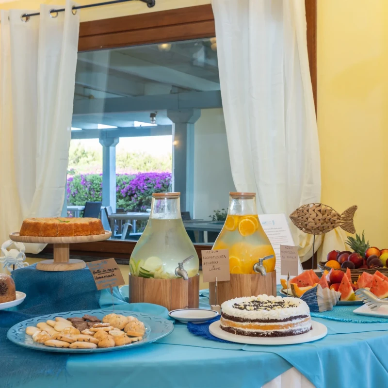 Tavolo della colazione con frutta fresca, dolci e succhi all'Hotel Luci del Faro con vista mare attraverso la finestra