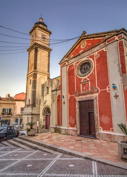 Chiesa con facciata rossa e campanile in pietra nella piazza principale di Calasetta al tramonto