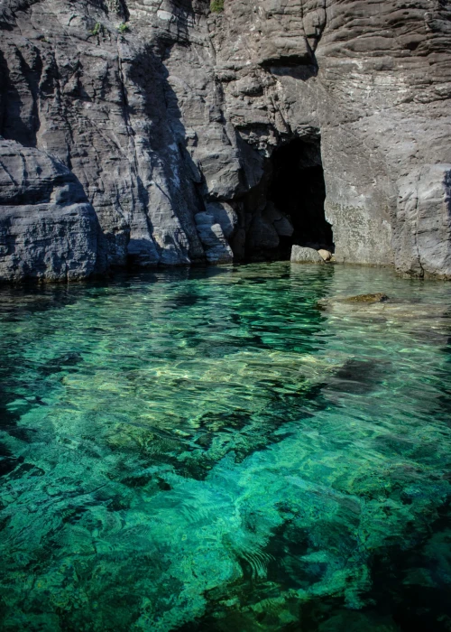 Acque turchesi cristalline con rocce stratificate grigie e grotta naturale sulla costa di Calasetta