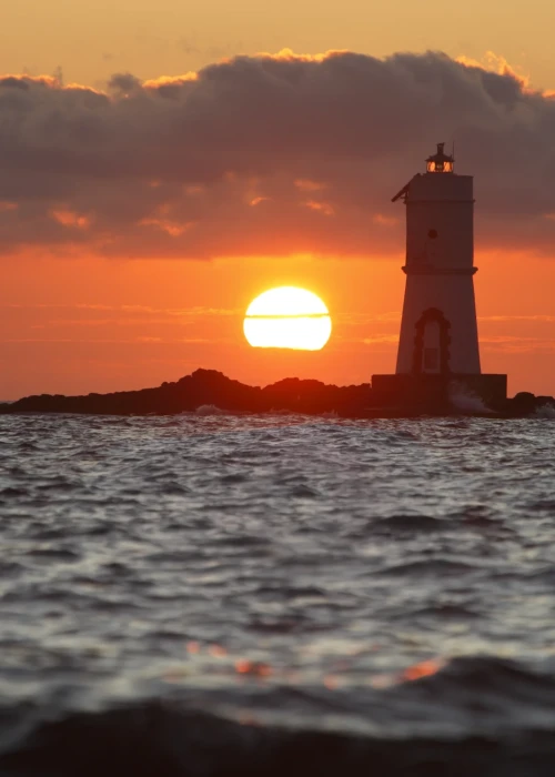 Tramonto sul mare con faro di Calasetta illuminato dal sole all'orizzonte, vista dalla spiaggia