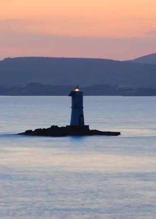 Faro isolato al tramonto sulle acque calme di Calasetta con colline all'orizzonte