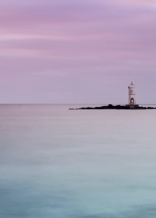 Faro solitario su isolotto roccioso al tramonto con mare calmo e cielo rosa-violaceo a Calasetta