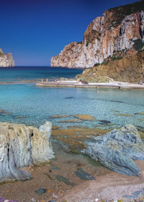 Spiaggia di Calasetta con acqua turchese trasparente, scogliere calcaree bianche e faraglione roccioso sullo sfondo