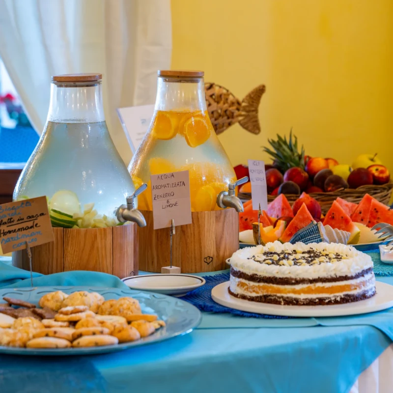 Buffet colazione con torta, frutta fresca, succhi e prodotti da forno su tavolo con tovaglia blu all'Hotel Luci del Faro