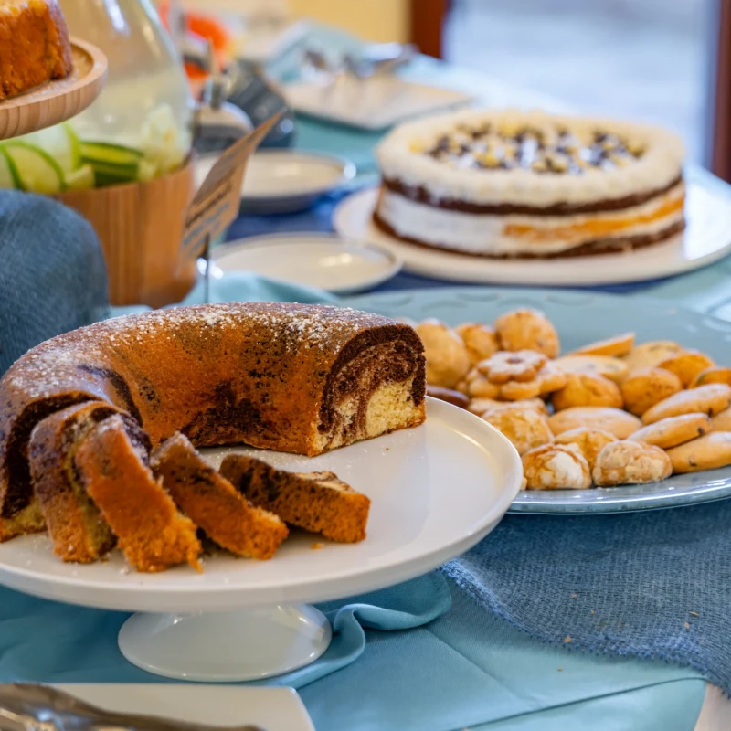 Tavolo da buffet con ciambellone al cioccolato, torte e biscotti su alzatine bianche con tovaglia azzurra