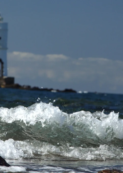 Onda che si infrange sulla costa di Calasetta con il faro bianco sullo sfondo e mare blu