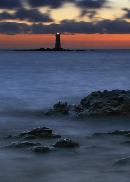 Faro di Calasetta al tramonto con mare calmo e scogli in primo piano sotto cielo arancione e blu
